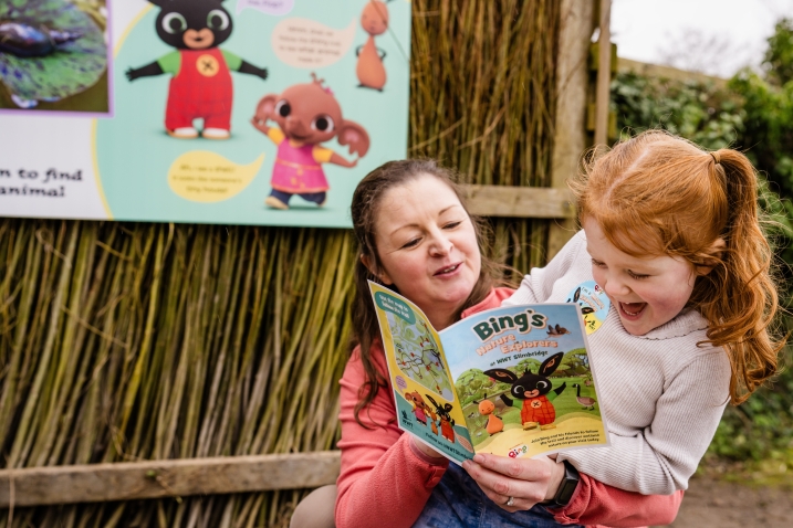A child sits on her mothers knee whilst holding a Bing activity booklet. In the background is a Bing board featuring the characters and information.